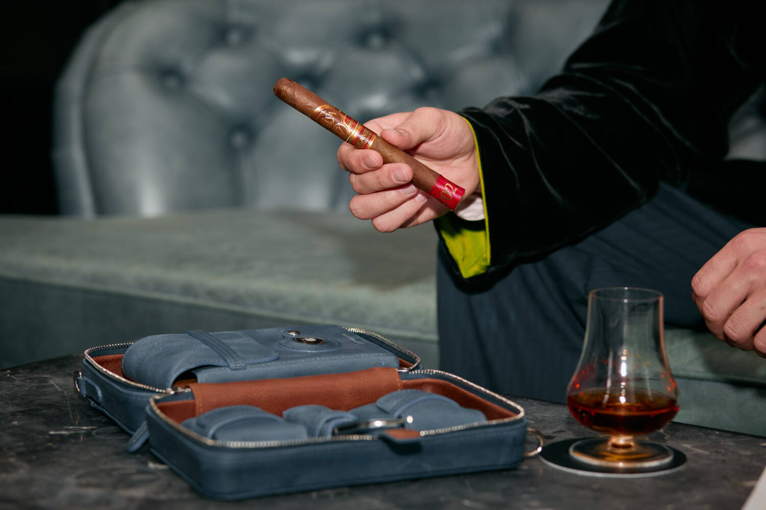 A person examining a fine cigar next to a leather cigar case and a glass of aged whiskey, signifying the luxury of cigar and drink pairings.