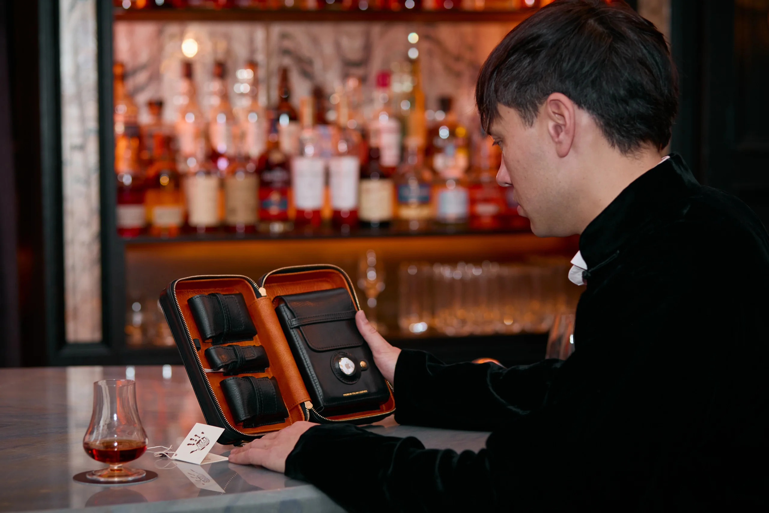 A cigar aficionado at a bar with a Britannia Nexus cigar case and a selection of fine spirits in the background.